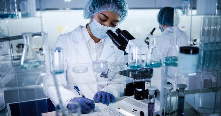 Multi ethnic, female team studying DNA mutations. Using microscope in protective workwear