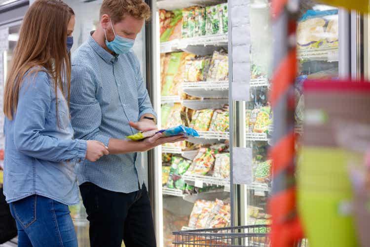 Young couple with protective face masks choosing frozen food in supermarket
