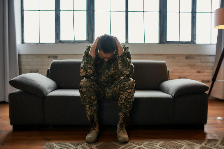 Constant pain. Full length shot of middle aged military man holding his head in pain and depression sitting on the couch. Soldier suffering from psychological trauma. PTSD concept