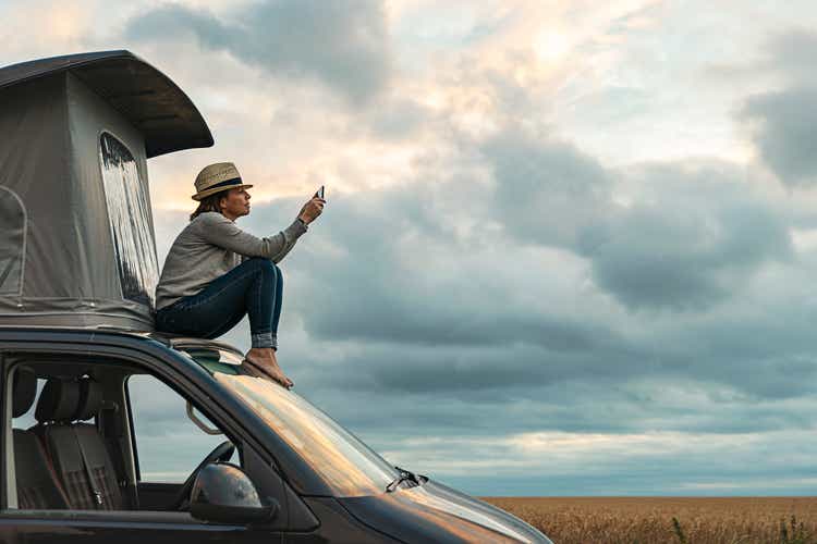 Woman taking a photo whilst sitting on roof of camper at sunset