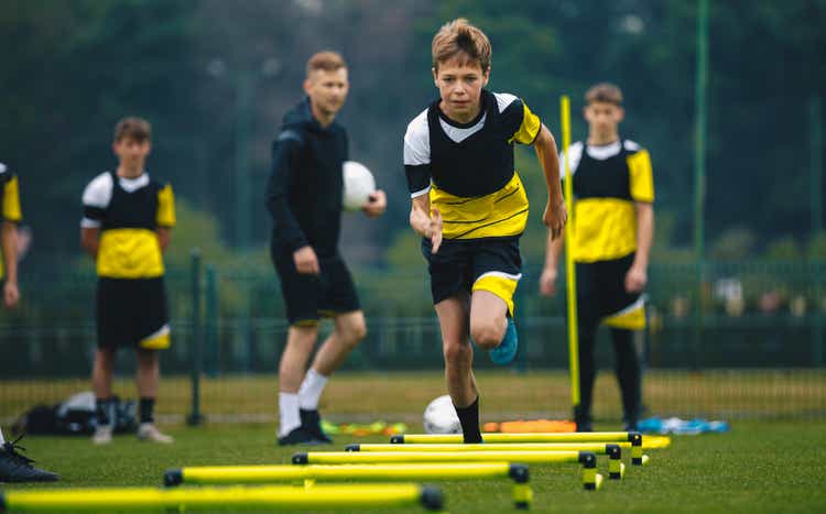 Teenager Boy Soccer Player in Training. Young Soccer Players at Practice Session with Coach. Footballer Jumping Over Hurdles. Boys Running Youth Agility Ladder Drills. Soccer Ladder Exercises