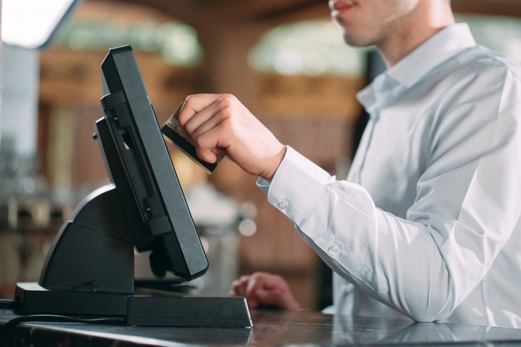 small business, people and service concept - happy man or waiter in apron at counter with cashbox working at bar or coffee shop.