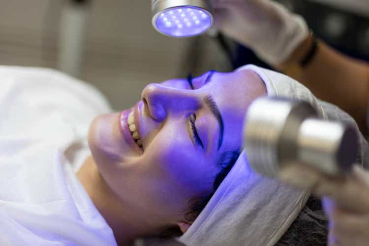Female Beautician Doing Blue Light Therapy On face of pretty young woman