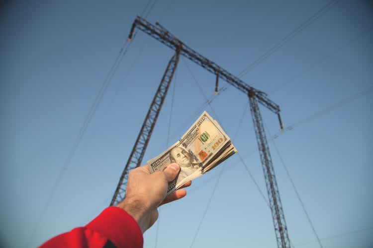 Male hand holding dollar banknotes on high voltage electric transmission tower background.