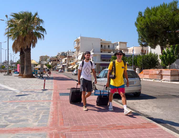 Two male tourists walk with suitcases and a tent along the promenade of the resort town on the Greek island Evia on a summer day