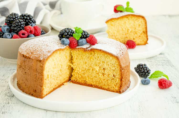 Homemade classic vanilla sponge cake or biscuit sprinkled with powdered sugar and fresh berries on top on a white plate on a light wooden background.
