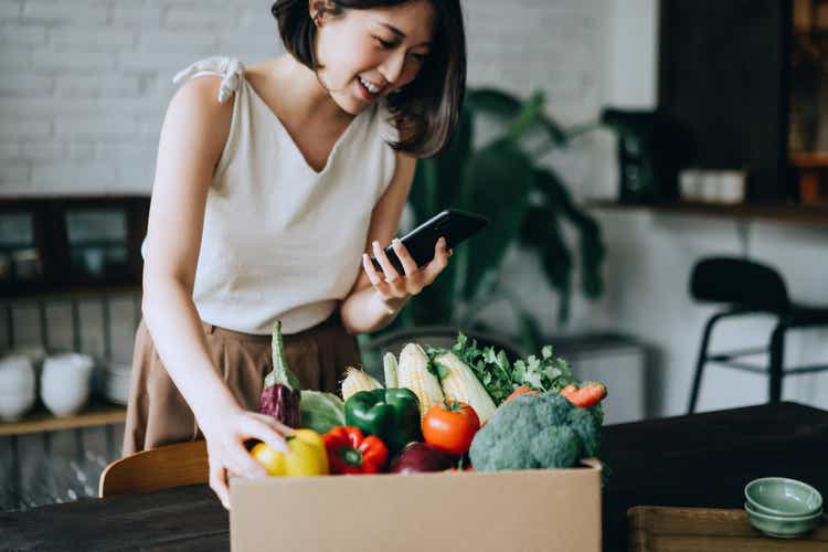 Beautiful smiling young Asian woman doing home delivery grocery shopping online with mobile app device on smartphone at home, with a box of colourful and fresh organic vegetables and fruits on the table