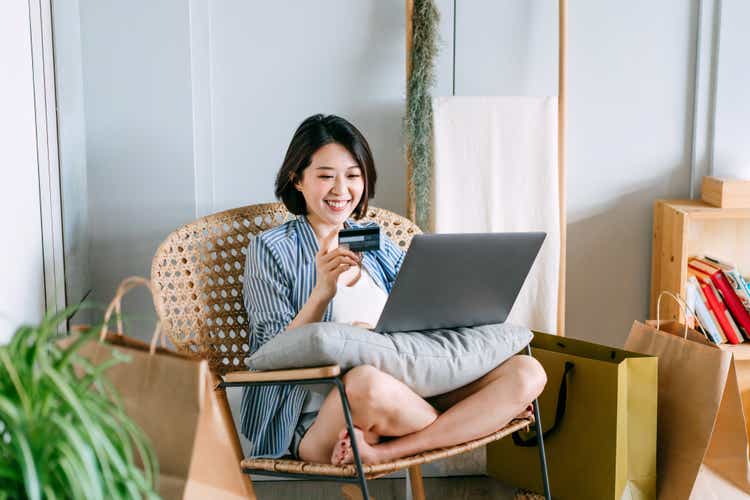 Beautiful smiling young Asian woman sitting on a rattan armchair in the living room at home, shopping online with laptop and making mobile payment with credit card. With some shopping bags next to her
