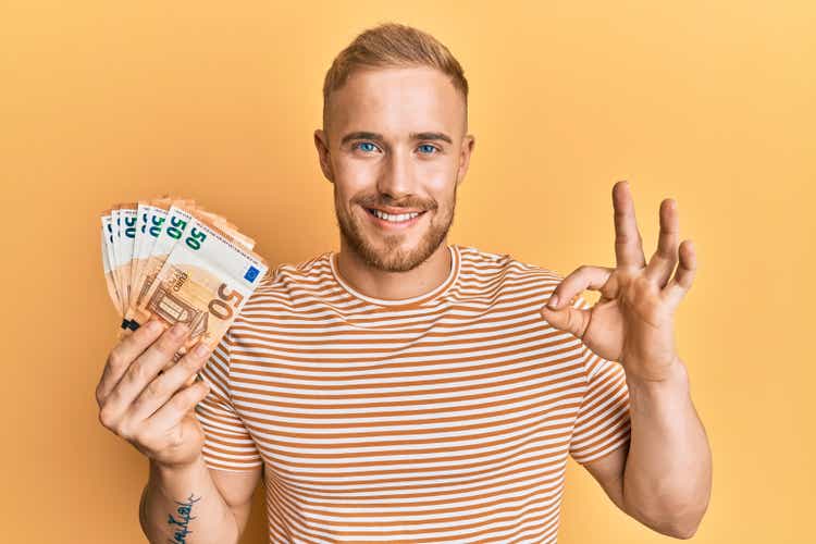 young caucasian man holding bunch of 50 euro banknotes making ok sign with fingers smiling friendly gesture thumbs up symbol