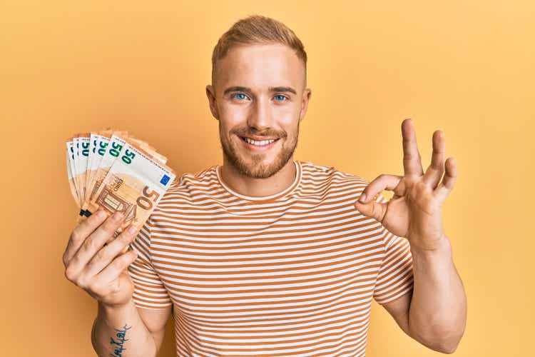 Young caucasian man holding bunch of 50 euro banknotes doing ok sign with fingers, smiling friendly gesturing excellent symbol