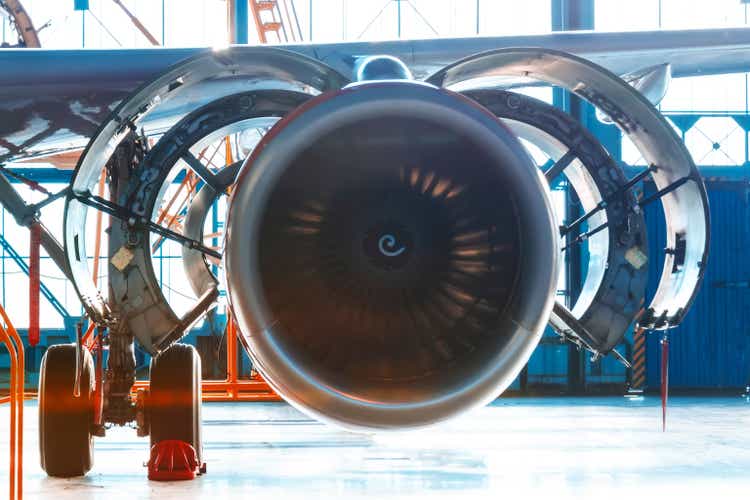 Airplane engine repair, hood flap open. View of wing with turbofan and landing gear. The bright light behind the aircraft hangar gates.