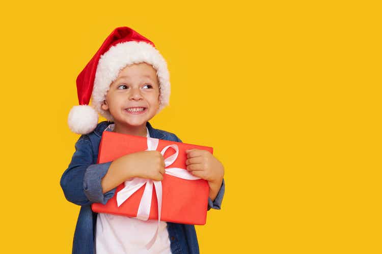 Portrait of a satisfied little child boy in christmas Santa hat. laughing isolated over yellow background. Holds a gift box. Preparing for the New Year holidays