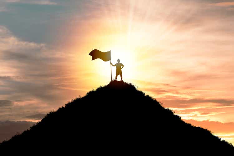 Business achievement goal and successful concept, Silhouette Man standing and holding flag on the top of the mountain with cloudy sky and sunlight.