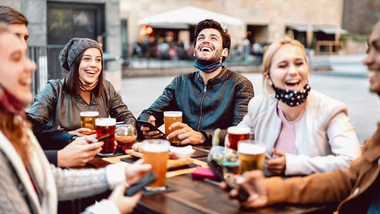Young friends drinking beer wearing face mask - New normal lifestyle concept with people having fun together talking on happy hour at outside brewery bar - Bright warm filter with focus on central guy