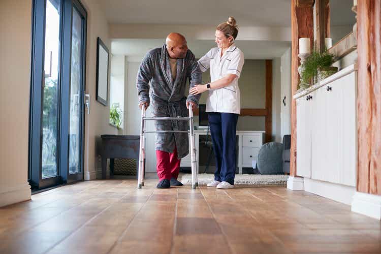 Senior Man In Dressing Gown Using Walking Frame Being Helped By Female Care Worker