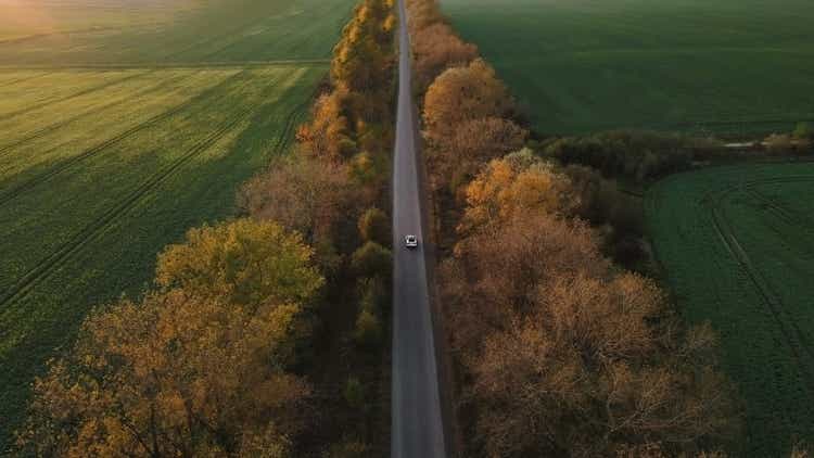 Aerial view Electric Car Driving on Country Road. Luxury modern vehicle riding fast along trees and fields. Cinematic drone shot flying over gravel road with trees at sunset