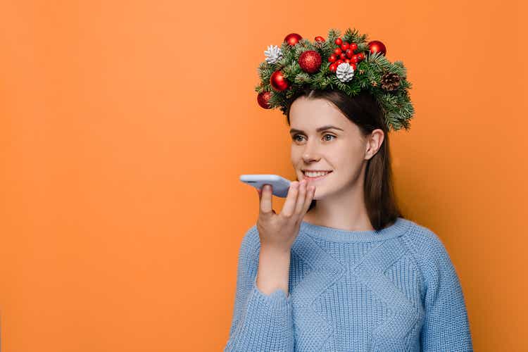 Translator app concept. Cute young woman holds phone talks on speakerphone with friend, makes voice recognition or request uses internet services through virtual assistant, wears Christmas wreath.