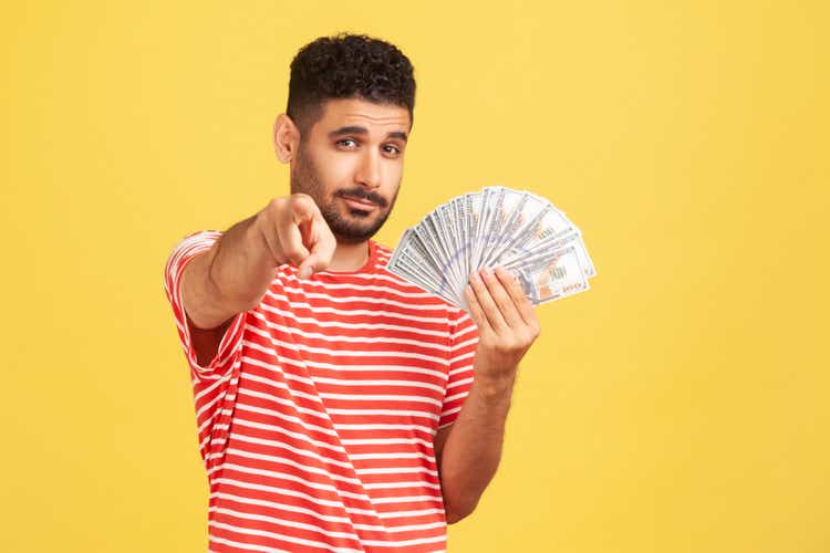 Self confident rich man with beard in striped t-shirt holding fan of dollars and pointing finger at camera with serious expression, easy profit.
