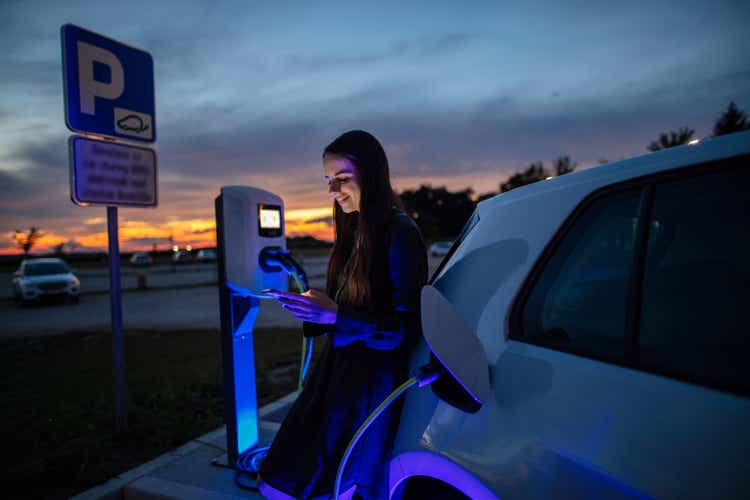Woman charging her electric car on gas station at night. Woman using mobile phone while waiting for electric car to charge in the parking lot at night