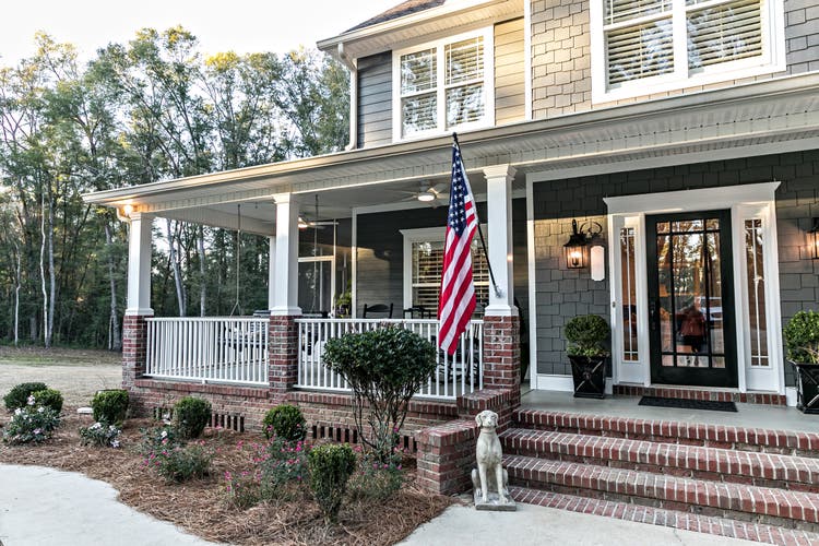 Front door entrance to a large two story blue gray house with wood and vinyl siding and a large American flag.