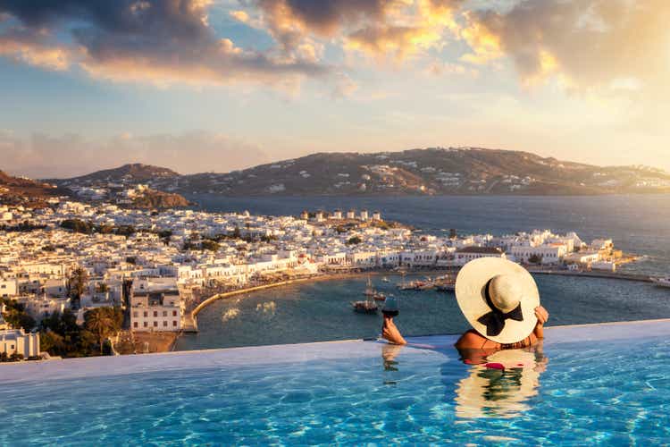 A woman with a glas of wine in a swimming pool enjoys the view over the town of Mykonos island, Greece