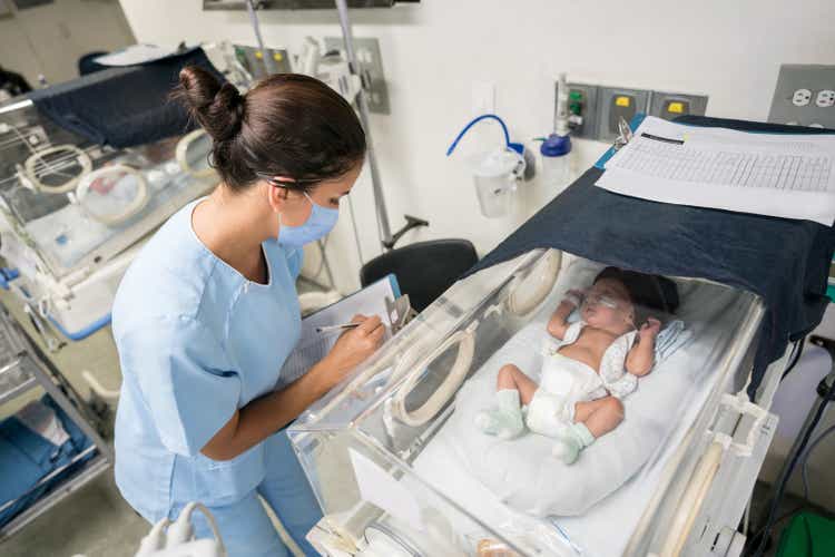 Nurse checking on a premature newborn in an incubator while wearing a facemask