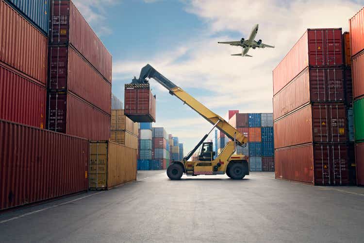 Airplane flying above container port.