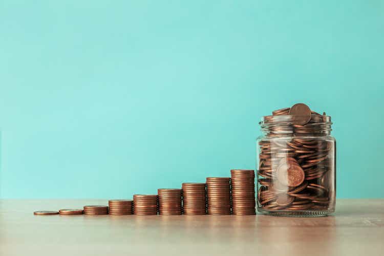 Stock photo of an ascending staircase of coins on a blue background with a jar full of coins. Saving concept