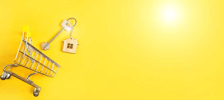 Sleutelhanger in de vorm van houten huis met sleutel op een gele achtergrond en boodschappenwagentje. Hypotheek, huur en aankoop onroerend goed, lente- en zomeraanbieding voor de bouw. Kopieerruimte