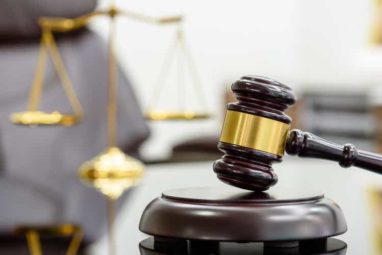 Wooden judge's gavel or wooden gavel and a soundboard used by a judge on a desk in a courtroom with a blurry brass scale of justice behind.