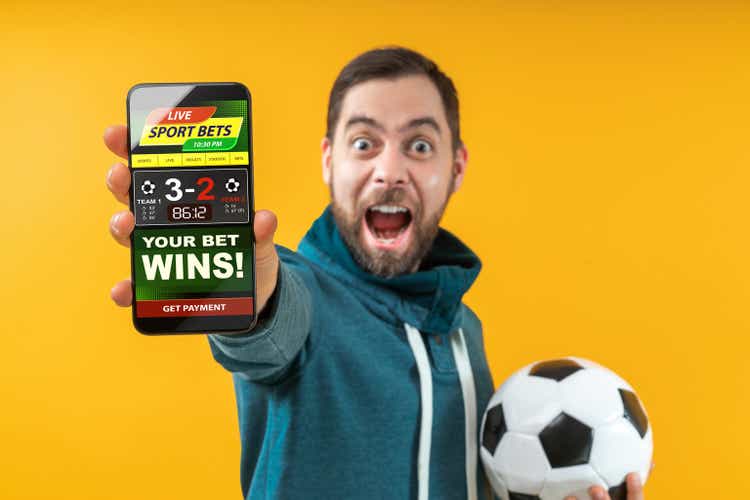 Happy excited football fan showing to the camera his smartphone with bookmaker"s website application interface template