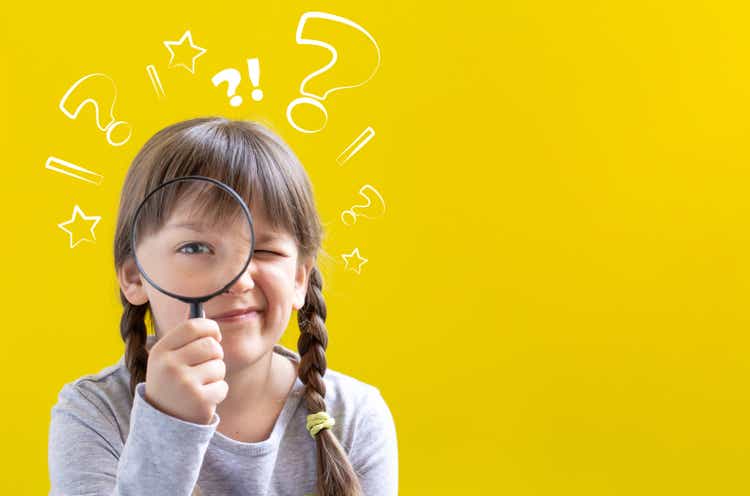 Cute suspicious little girl looking through a magnifying glass on yellow background with copy space.