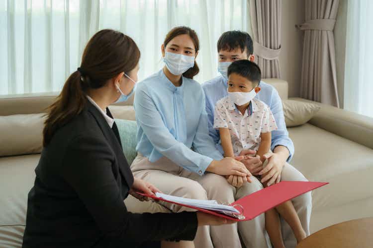 Asian woman"s insurance broker is offering details of health insurance coverage for COVID-19 to Asian family with father, mother and son in their living rooms at home.