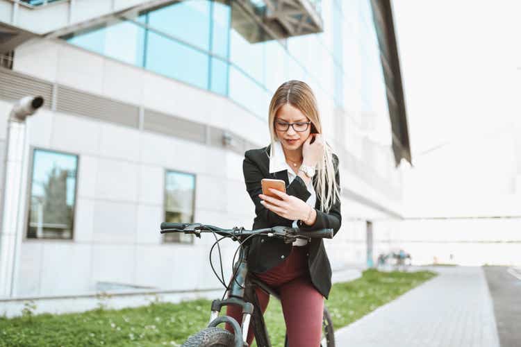 Smiling Beautiful Businesswoman Using Smartphone While Riding Bicycle Outside
