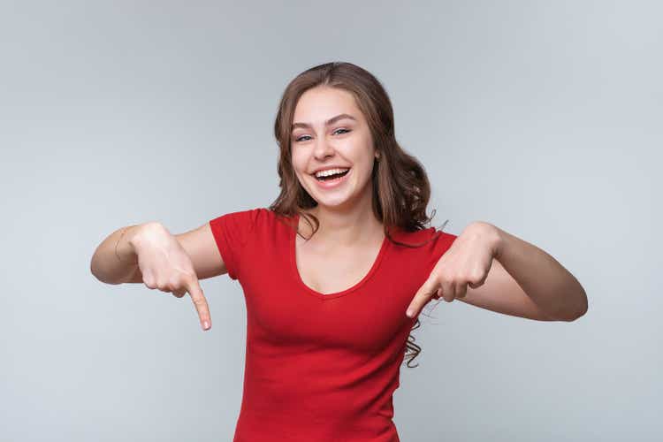 Happy young brunette woman smiling and pointing fingers down at your promo text, standing over gray background
