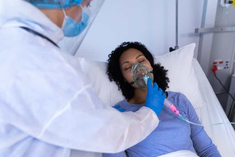 Caucasian doctor in ppe suit checking on female patient in hospital bed with oxygen ventilator