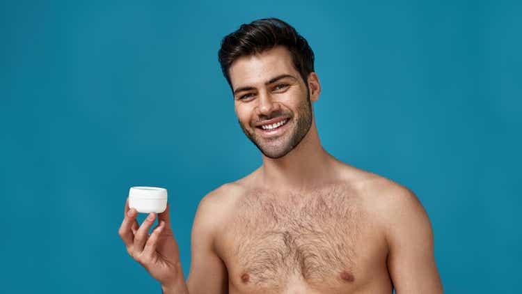 Portrait of cheerful naked brunette guy holding white jar with effective cream for face, smiling at camera isolated over blue background