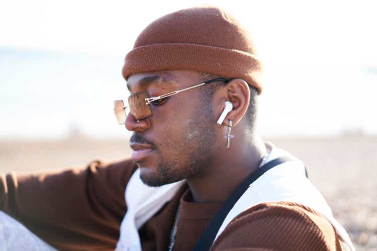 Stylish young man spending some time sitting on the beach listening to music with wireless earphones