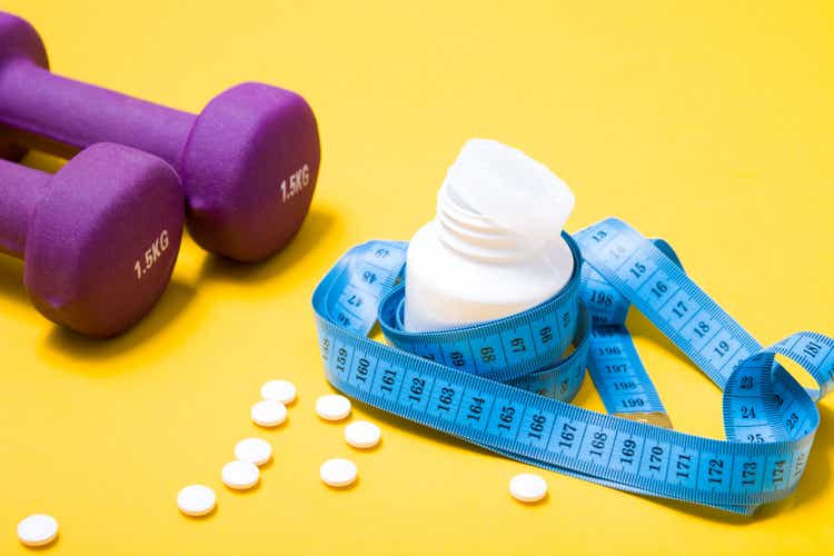 blue measuring tape around a white jar of tablets, tablets are scattered on a yellow background, dumbbells of purple color, copy space, slimming concept