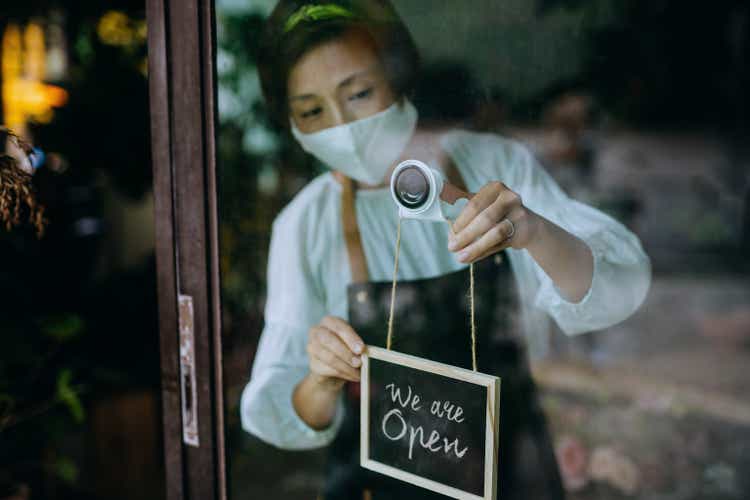 Asian female florist, owner of small business flower shop wearing protective face mask, starting her business day. Hanging the open sign on the door at the shop. New normal of everyday business practice