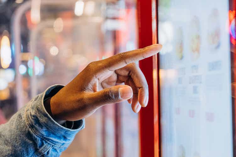 A close-up finger selects a burger on an electronic touch screen at a self-service checkout at a fast food restaurant