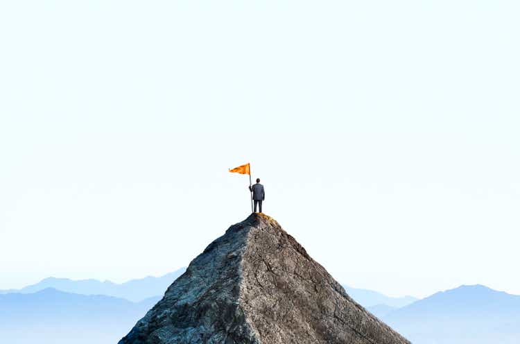 Businessman At Top Of Mountain Peak Holds Large Flag