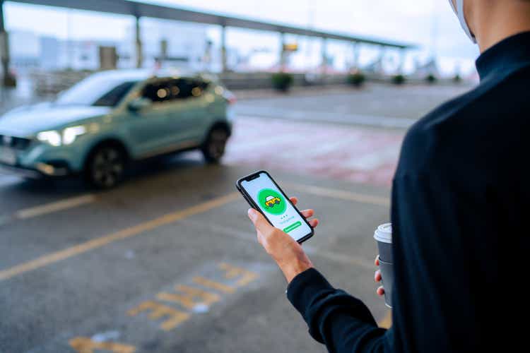 Chic young Asian man using mobile app device on smartphone to order a taxi pick up service by the urban road in downtown city street. Speedy and trustworthy service. Carsharing and business on the go themes