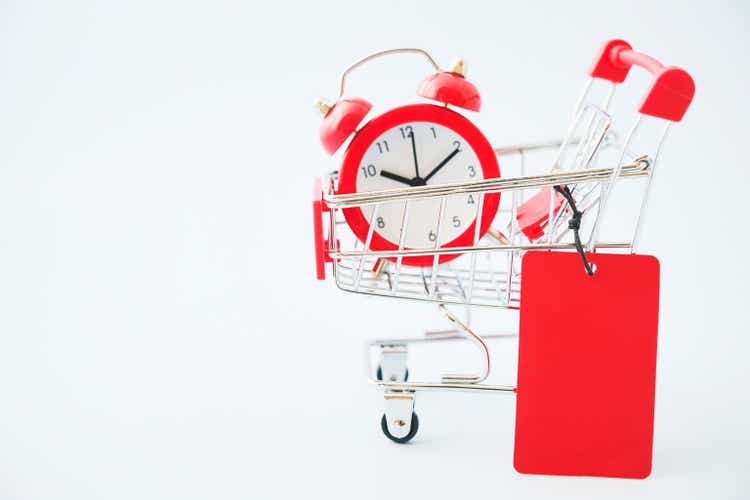 red paper price tag , shopping cart and blurred analog alarm clock on light blue background for sale , promotion concept