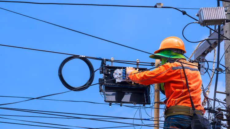 technician on wooden ladder is working to install fiber optic and splitter box on electric pole against blue sky