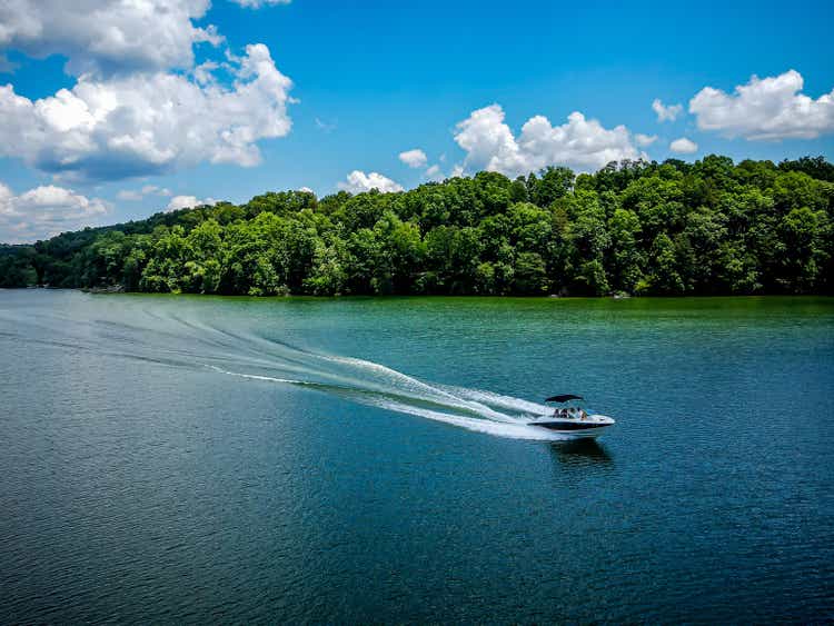 Boat racing across a lake with wake behind it, green trees in the background, and clouds in the sky