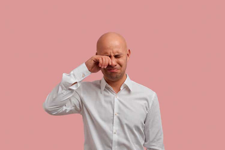 Unhappy sad bald man with bristle rubs tears wants to cry feels desperate has misunderstandings and bad conversation with people. Dressed in white shirt, isolated over pink background.