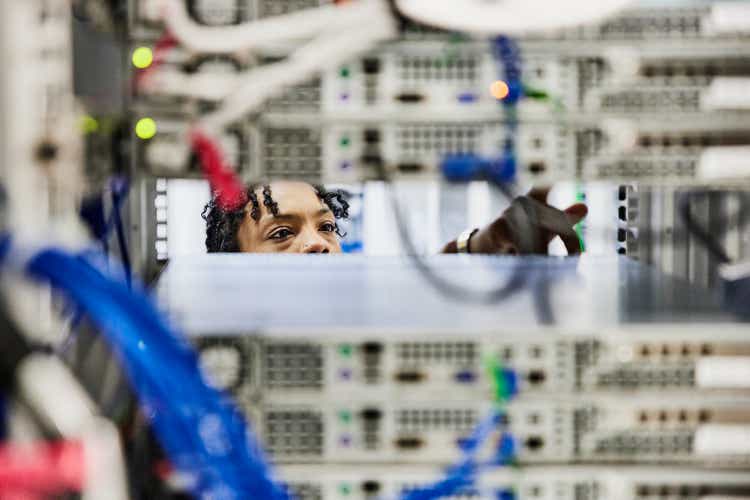 Medium shot looking through server rack of female IT professional working in data center