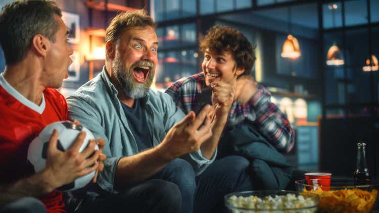 Night at Home: Three Soccer Fans Sitting on a Couch Watch Game on TV, Use Smartphone App to Online Bet, Celebrate Victory when Sports Team Wins. Friends Cheer Eat Snacks, Watch Football Play.