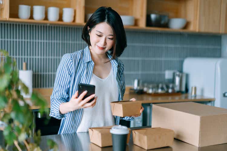 Beautiful smiling young Asian woman standing by the kitchen counter, ordering takeaway food using mobile app device on smartphone for a food delivery service. Technology makes life so much easier